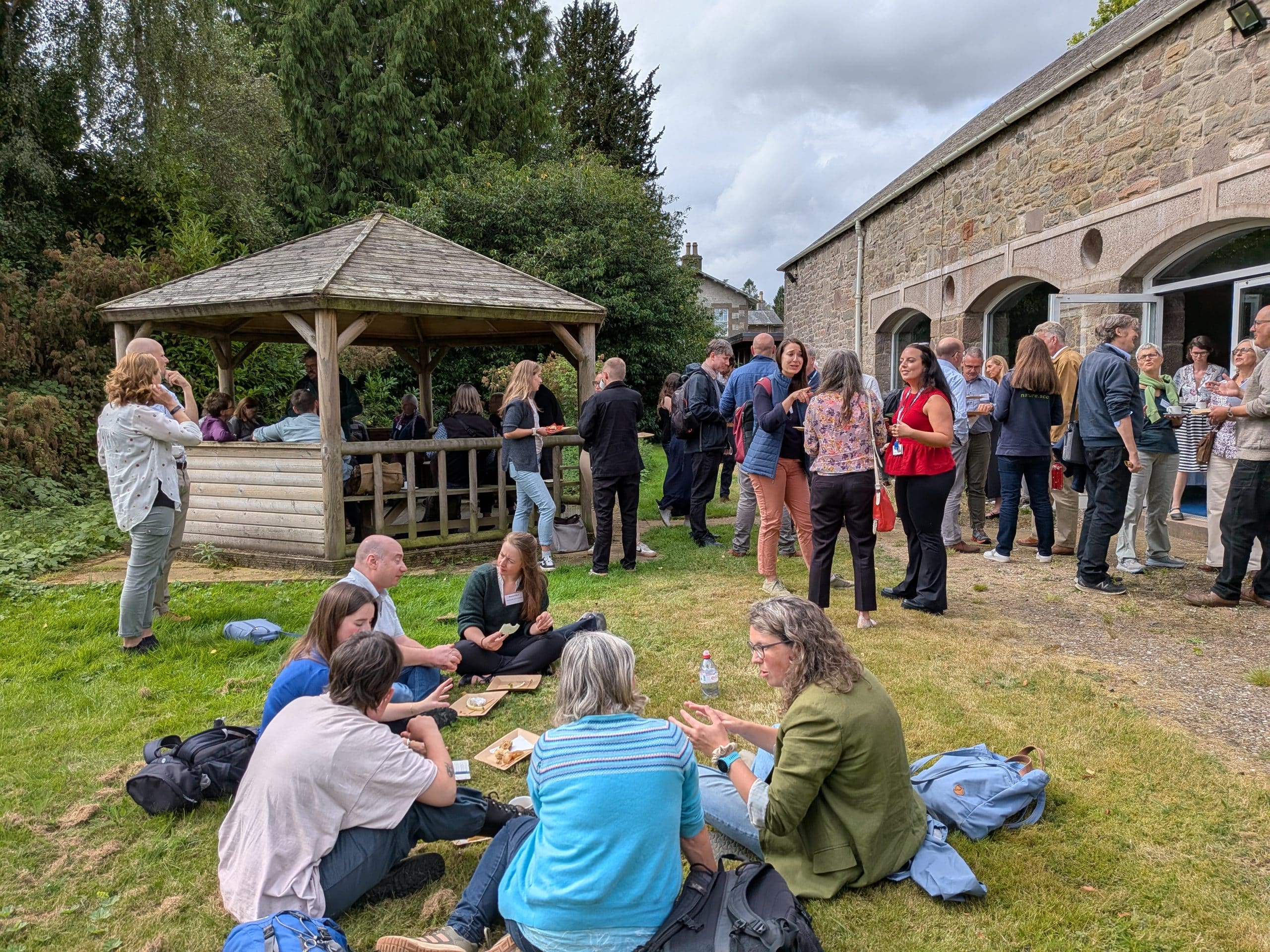 people eating in small groups, with some sitting on grass in the foreground and other standing on grass in the background